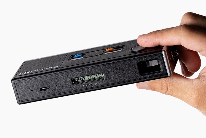 Hand holding the Camp Snap 110D, showing the small segmented LCD and USB C port on the back