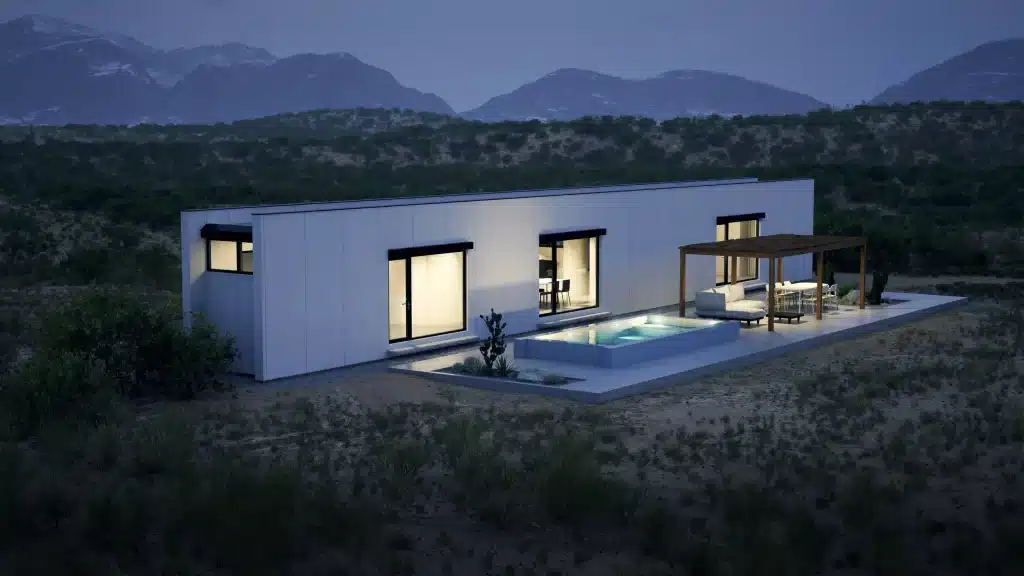 Monocabin XL5 prefab house at dusk with lit windows, a small pool and mountains in the background