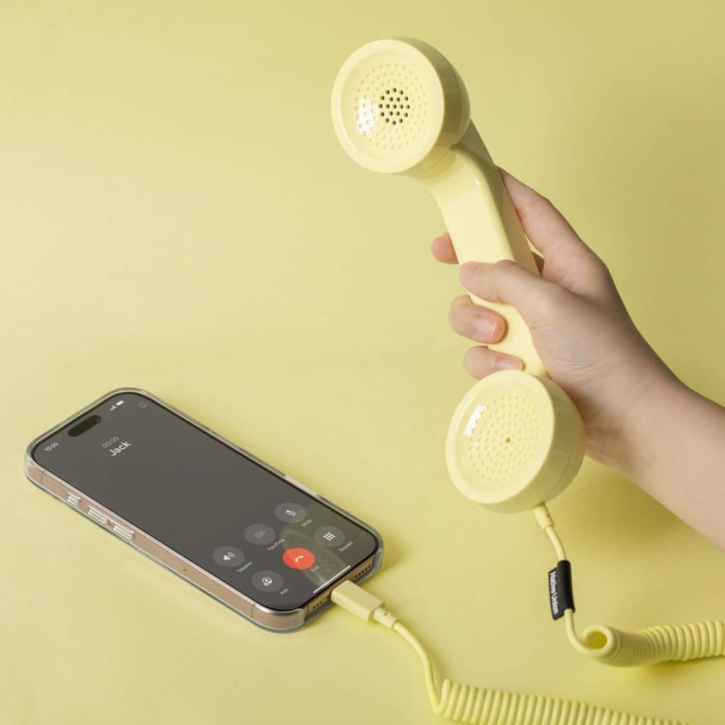 Hand holding a yellow Native Union POP Phone handset connected by coiled cable to a smartphone on a yellow background