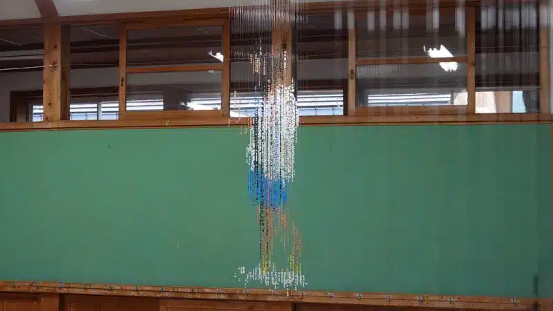 A child made of suspended beads standing against the green wall of an empty classroom